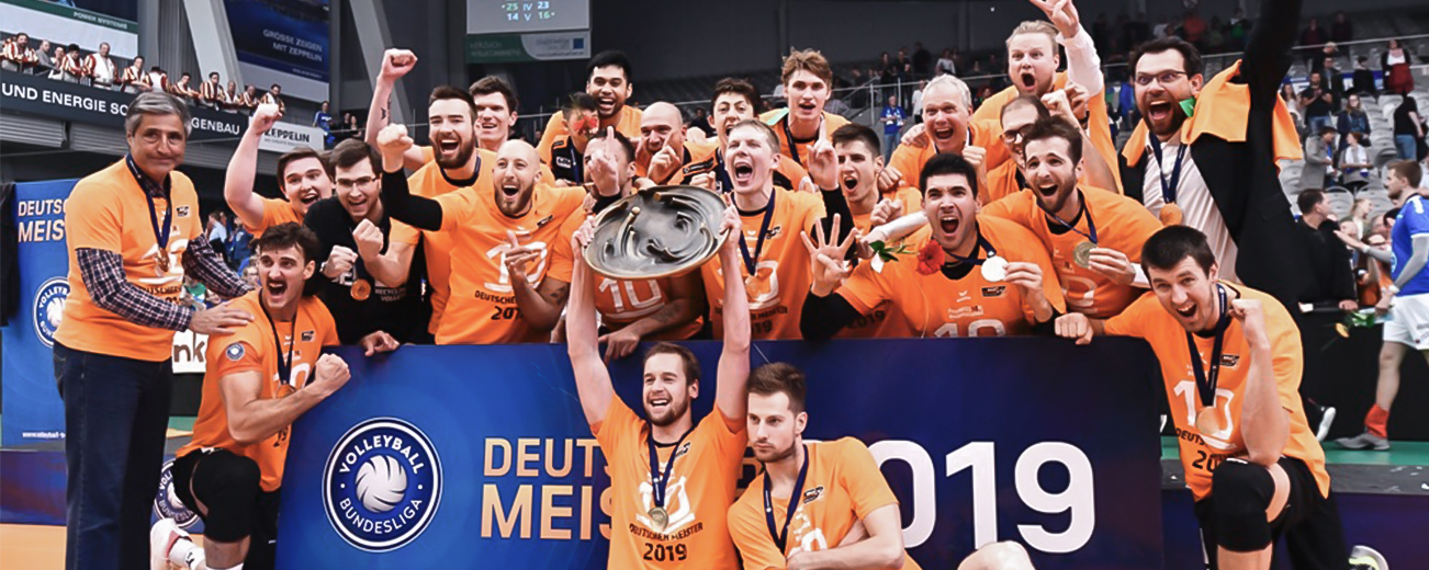 Gruppenfoto der Berlin Recycling Volleys zur Siegerehrung der deutschen Meisterschaft 2019, mit einem Transparent, mehreren Trainern sowie einer Trophäe in den Händen eines Spielers